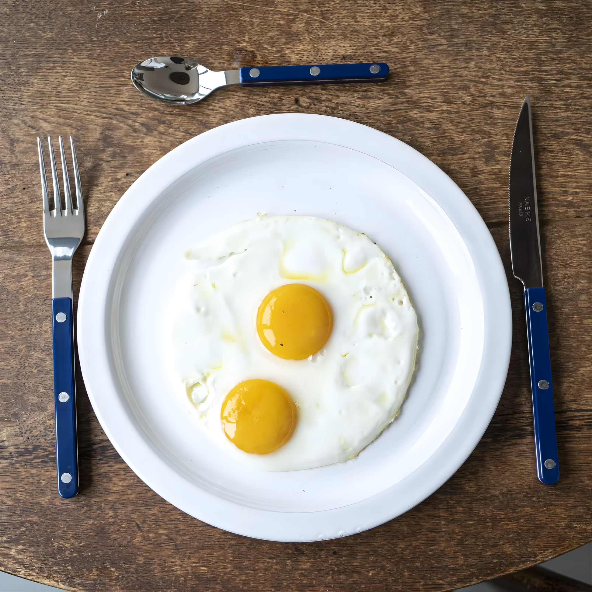 Shown with a knife and teaspoon beside a plate, the Sabre Bistrot Cutlery Fork 22cm Navy Blue which is made from Inox 18/10 Manche, stainless steel designed to be both traditional and contemporary, dishwasher safe at 113°F (45°C) maximum, not suitable for microwave oven.
