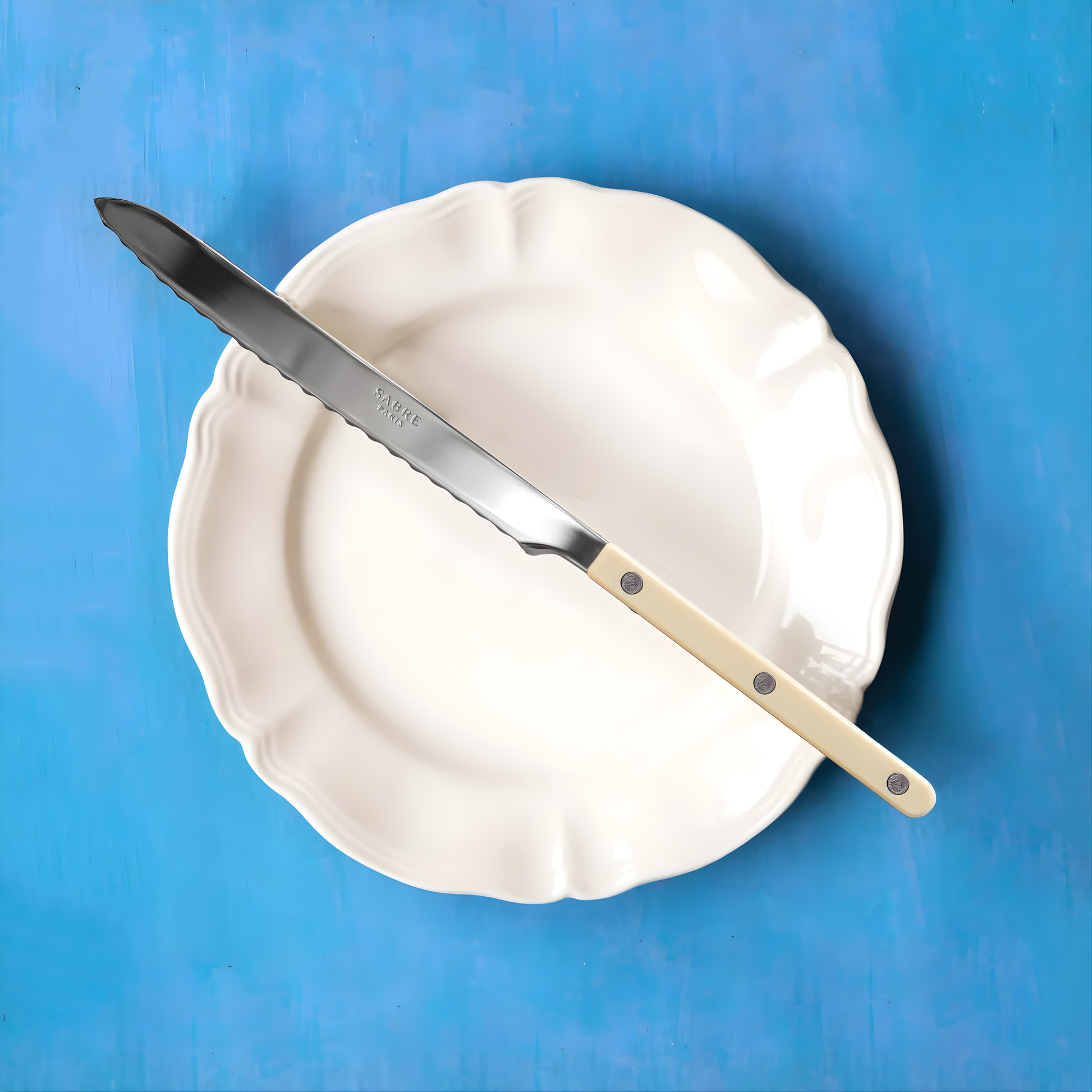 Sabre Bistrot Cutlery Ivory Breadknife on a plate, it has a long serrated blade to cut all types and sizes of bread, pastries, sweet and savoury cakes.