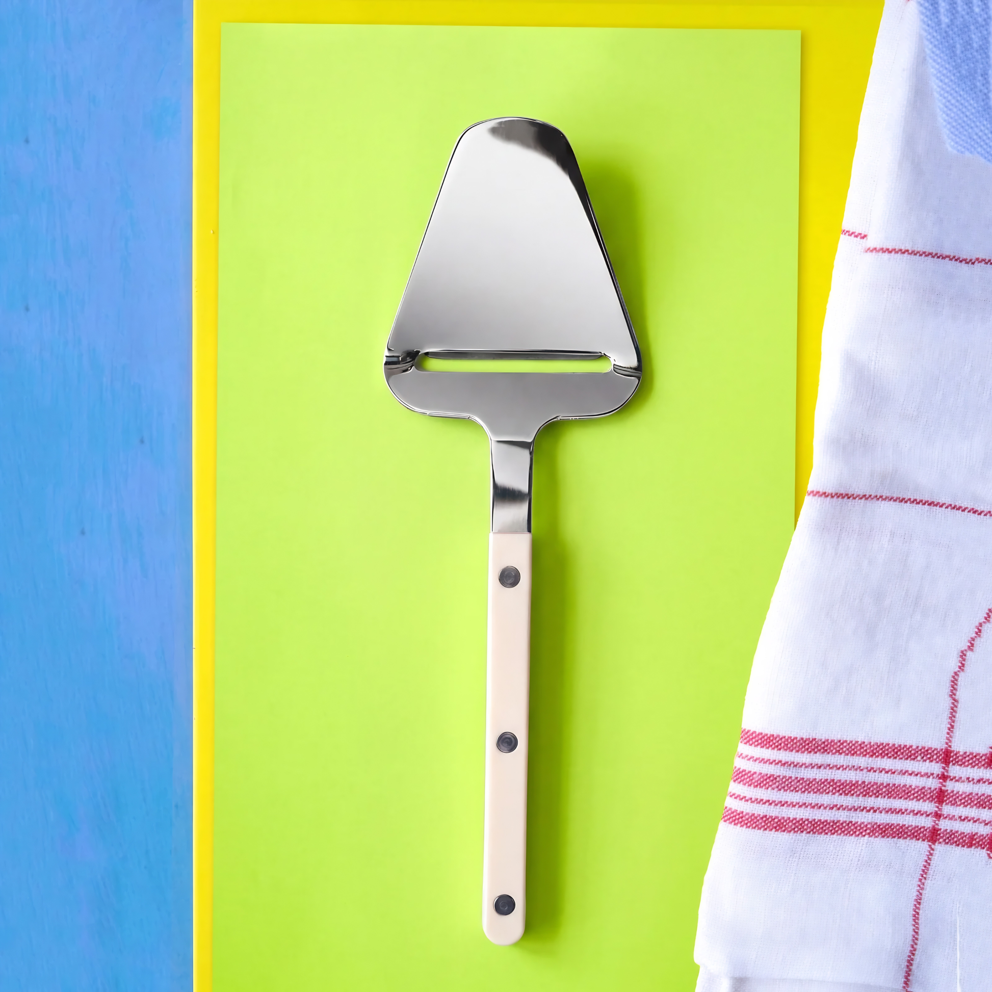 Sabre Bistrot Cutlery Ivory Cheese Slicer on a green paper surface, the slicer has rivets visible and can create thin shavings of cheeses.