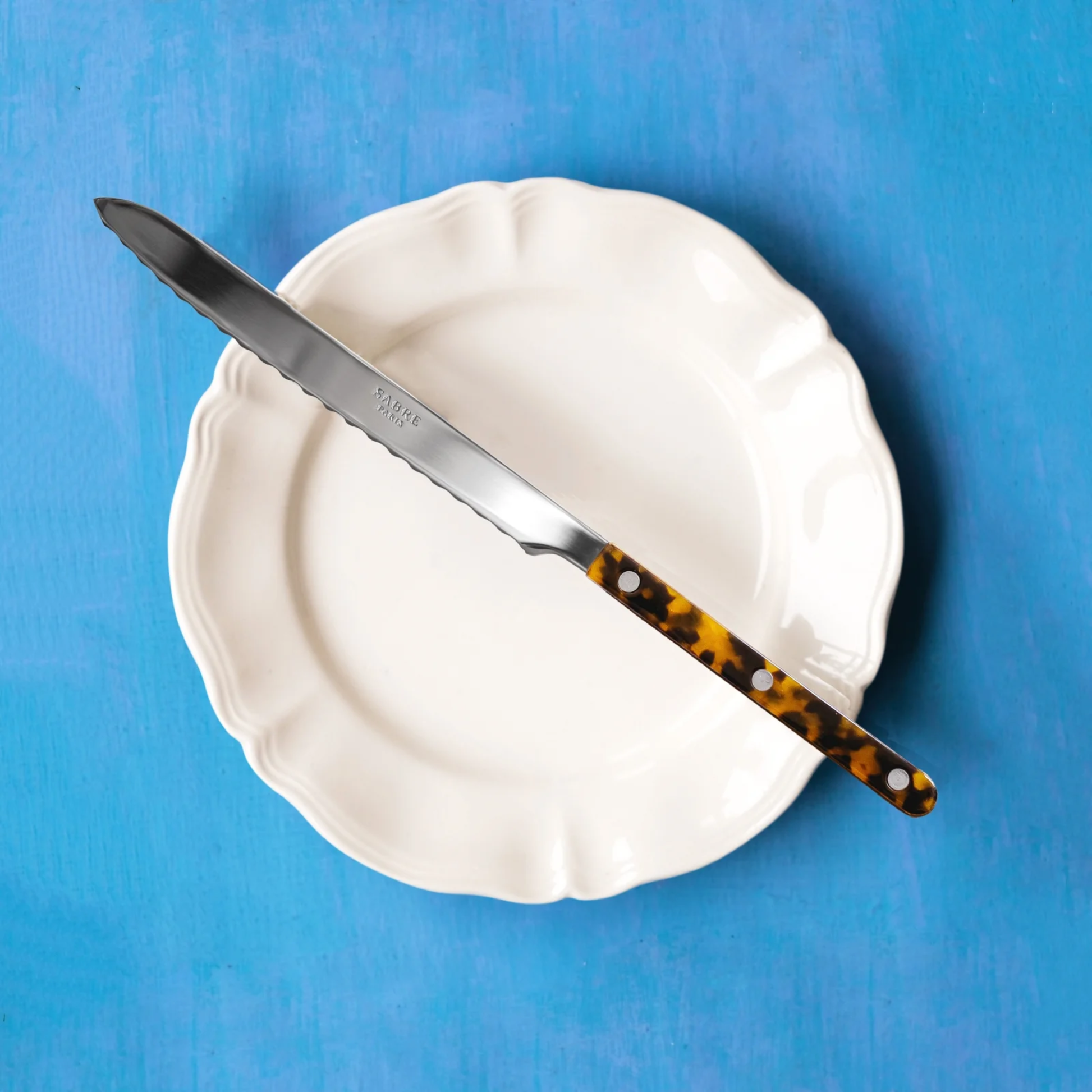 Sabre Bistrot Cutlery Tortoise Breadknife on a plate, it has a long serrated blade to cut all types and sizes of bread, pastries, sweet and savoury cakes.