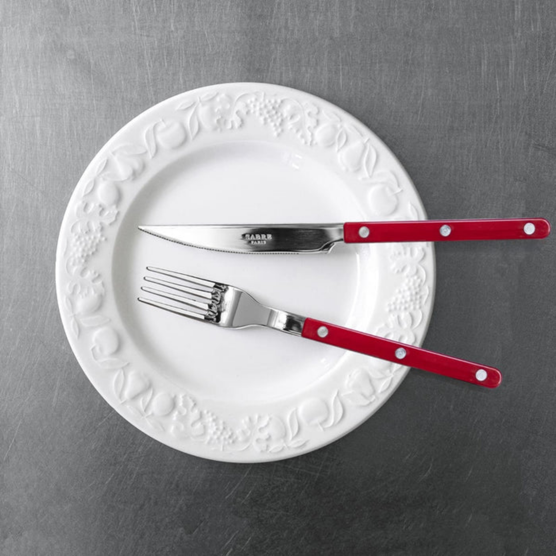 On a plate with a knife Sabre Paris Bistrot Dinner Fork Burgundy - 22cm stainless steel fork with colorful nylon handle and visible rivets. Inspired by French cafés, this elegant and durable fork blends classic and modern design, available in gloss or vintage finishes and dishwasher-safe.