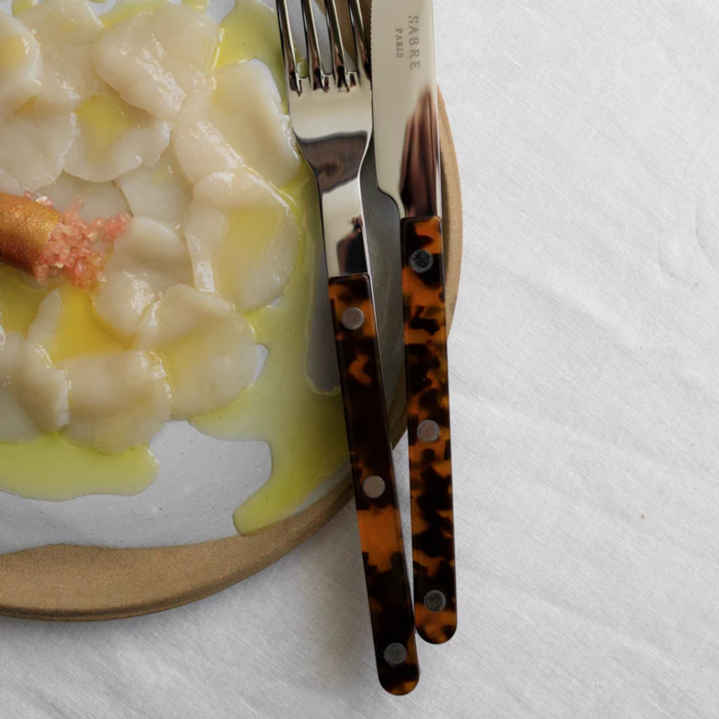 Close up showing the logo with food beside Sabre Paris Bistrot Dinner Knife - 24cm stainless steel knife with colorful acrylic handle and café-style rivets. A sharp, elegant design blending traditional French charm with modern flair, perfect for everyday dining or refined table settings.