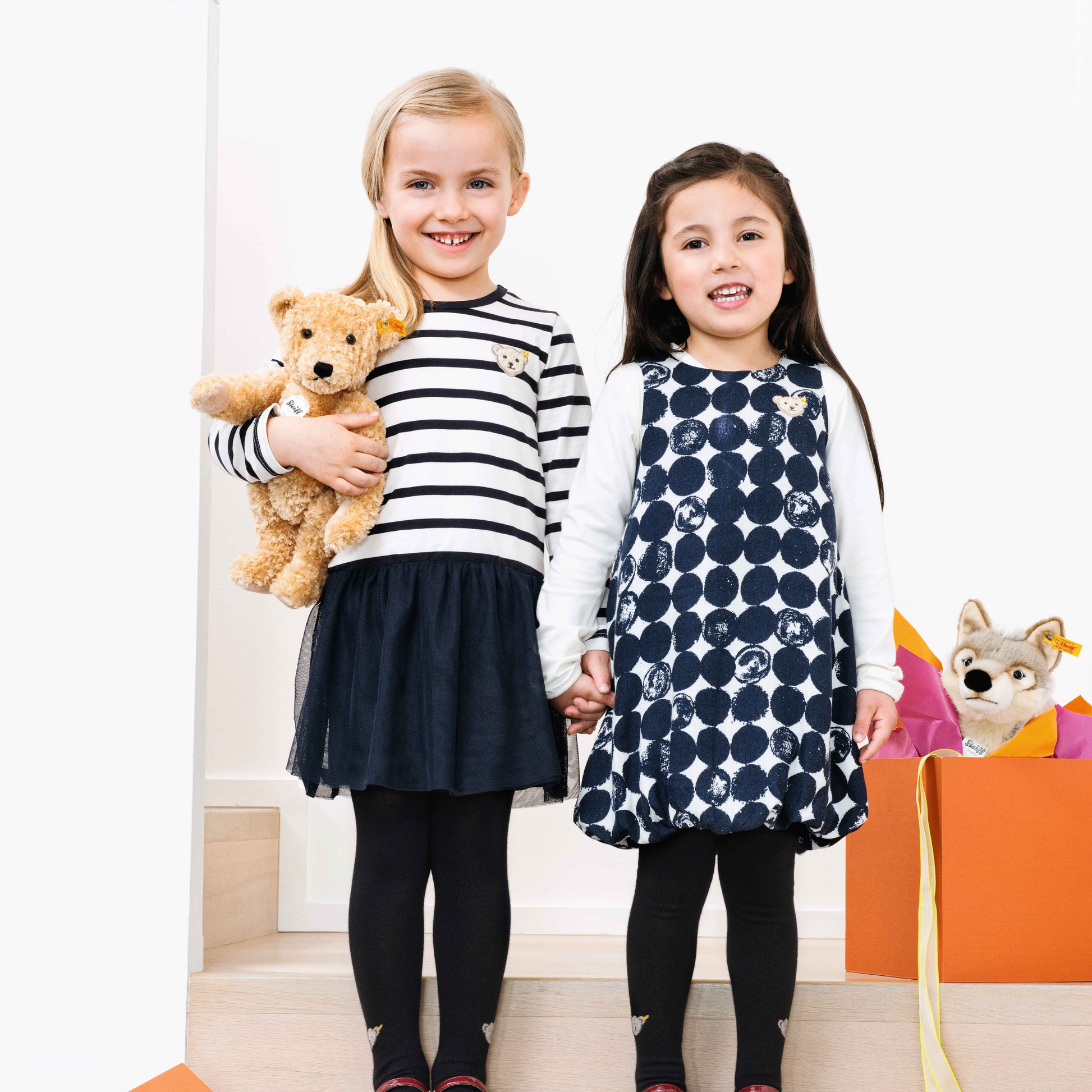 Front view of two girls with the Steiff Classic Teddy Bear Elmar 32cm Beige which is made from from cuddly, durable synthetic polyester with a five-way jointed, softly stuffed body for extra hugs. Features handmade shoe-button eyes, is machine washable, and carries the Steiff Button in Ear trademark of quality.