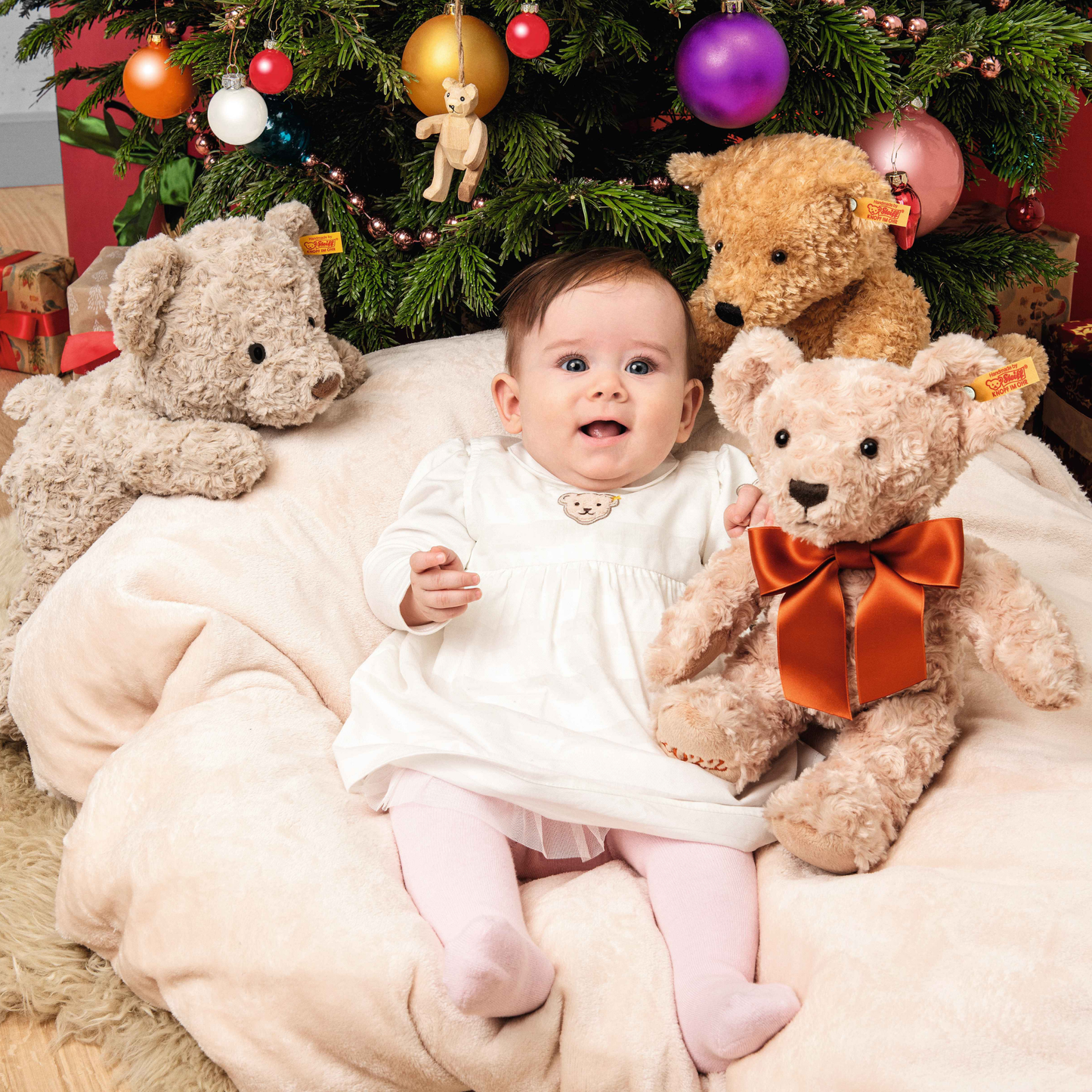 A baby girl on a bean bag chair with three types of Steiff Teddy bear including the Steiff Classic Teddy Bear Elmar 32cm Beige which is made from from cuddly, durable synthetic polyester with a five-way jointed, softly stuffed body for extra hugs. Features handmade shoe-button eyes, is machine washable, and carries the Steiff Button in Ear trademark of quality.
