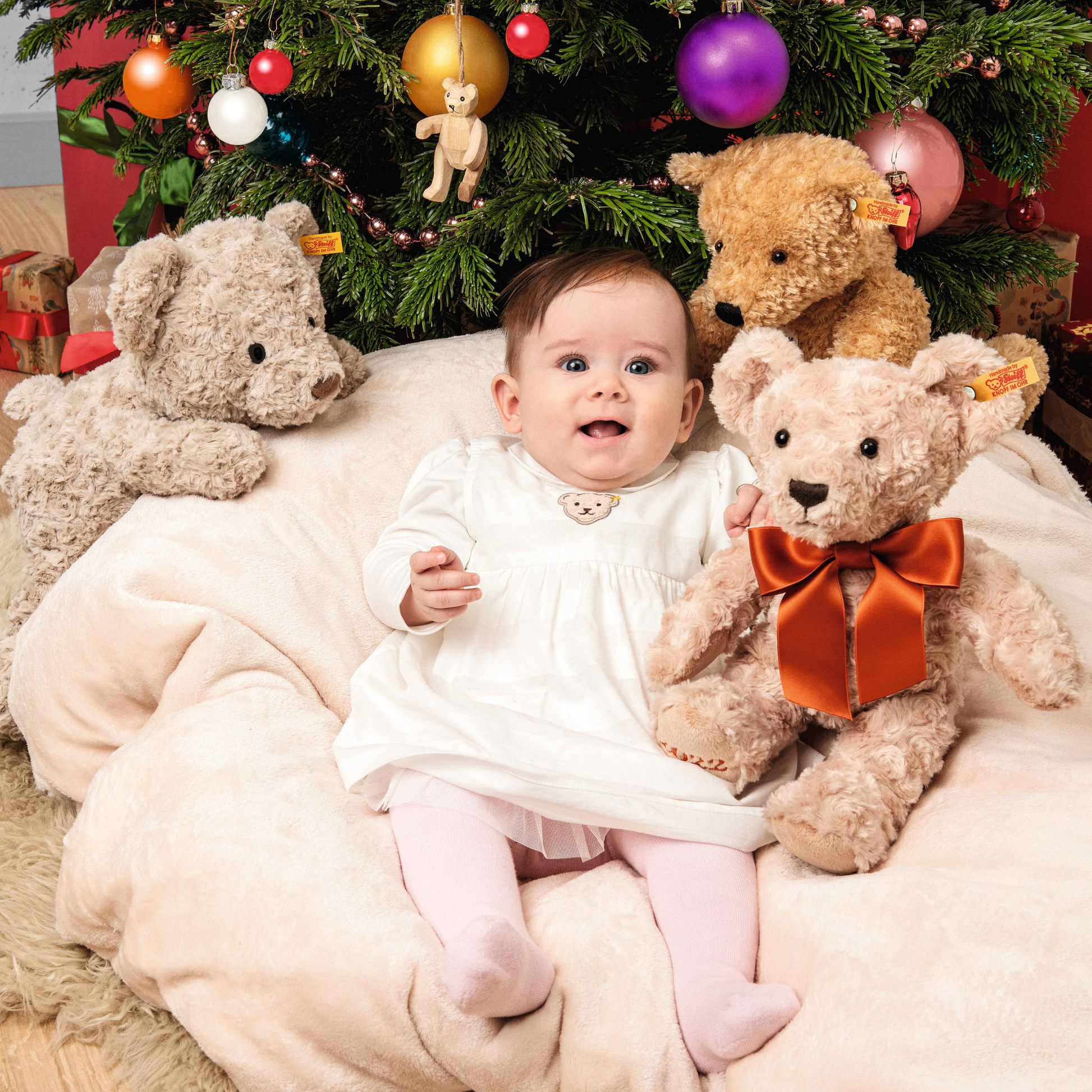 A baby girl on a bean bag chair with three types of Steiff Teddy bear including the Steiff Classic Teddy Bear Elmar 32cm Beige which is made from from cuddly, durable synthetic polyester with a five-way jointed, softly stuffed body for extra hugs. Features handmade shoe-button eyes, is machine washable, and carries the Steiff Button in Ear trademark of quality.
