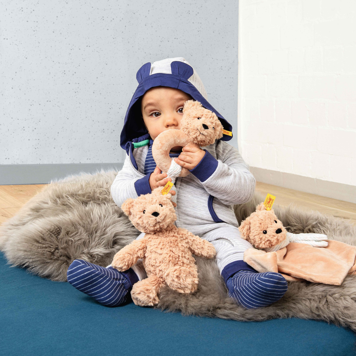 A baby girl playing with three teddy bear including the Steiff Classic Teddy Bear Jimmy 20cm Light brown which is made from from cuddly, durable synthetic polyester with a five-way jointed, softly stuffed body for extra hugs. Features handmade shoe-button eyes, hunchback inspired from a real bear, is machine washable, and carries the Steiff Button in Ear trademark of quality.
