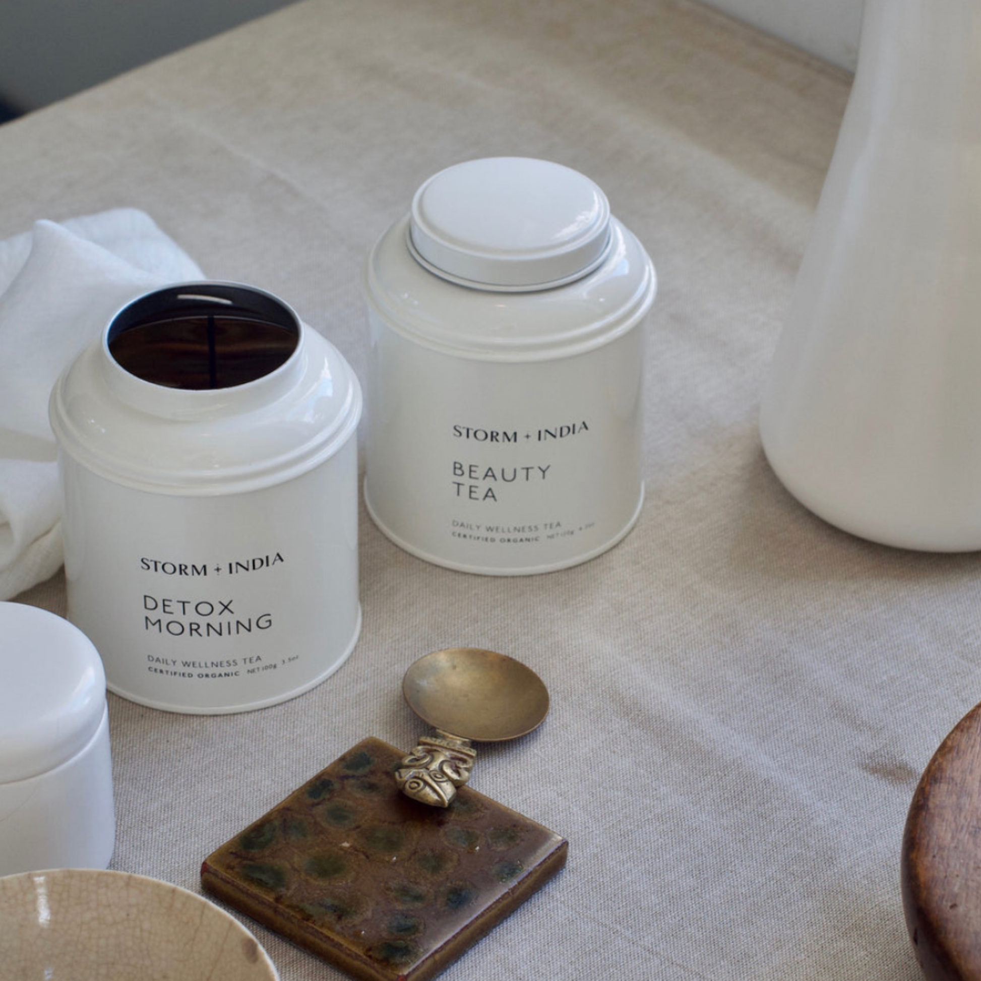 Storm and India Loose Leaf Tea in Tin 100g Detox Morning with lid open and Storm and India Beauty Tea in Tin sibe by side on linen covered table with tea accessories 