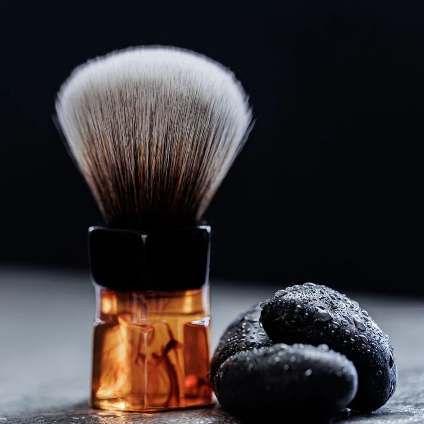 Shown beside small stones Yaqi Moka Express Synthetic Shaving Brush - Resin shaving brush with 24mm synthetic Mew Brown knot and palm-grip handle - Durable resin build with 52mm loft and 54mm handle height - Stylish smokey swirl design for smooth lathering and exfoliating shave experience